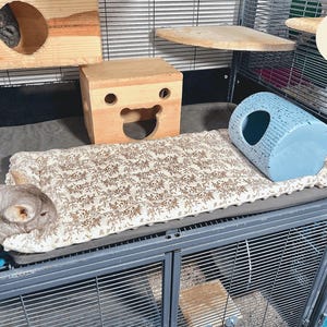 May include: A multi-level pet cage with wooden structures, a patterned bed, and a blue hideaway. The cage includes a chinchilla resting on the bed. The cage is made of metal and has multiple levels for the pet.