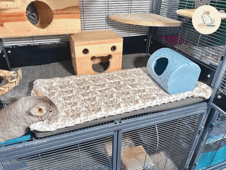 May include: A multi-level pet cage with wooden structures, a patterned bed, and a blue hideaway. The cage includes a chinchilla resting on the bed. The cage is made of metal and has multiple levels for the pet.