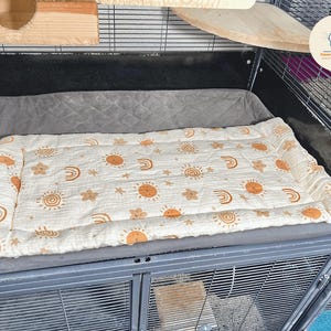 May include: A rectangular pet bed with a cream-colored top featuring a repeating pattern of orange suns, rainbows, and stars. The bed is placed inside a metal cage, resting on a gray cushion. The cage has wooden shelves.