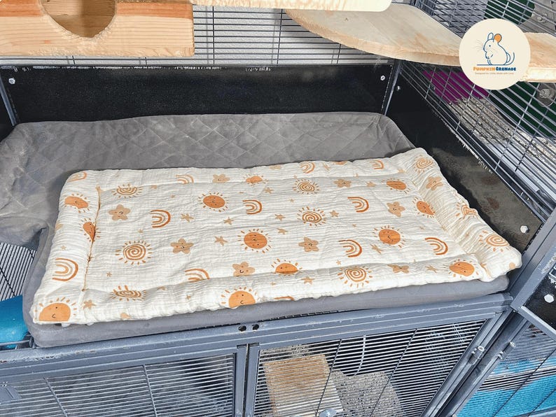 May include: A rectangular pet bed with a cream-colored top featuring a repeating pattern of orange suns, rainbows, and stars. The bed is placed inside a metal cage, resting on a gray cushion. The cage has wooden shelves.