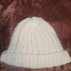 Könnte beinhalten: Eine cremefarbene, handgestrickte Beanie-Mütze. Die Mütze hat einen gerippten Saum und ein strukturiertes, engmaschiges Muster über die gesamte Krone. Die Mütze ist aus einem weichen, warmen Garn gefertigt.