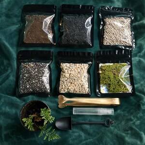 Puede incluir: Un conjunto de suministros de jardiner&iacute;a, que incluye tierra, guijarros y musgo en bolsas negras con cierre zip. Tambi&eacute;n se incluyen una peque&ntilde;a pala negra, pinzas doradas, un pulverizador y una maceta peque&ntilde;a con un helecho.
