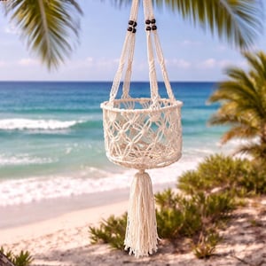 Pu&ograve; includere: Un portavaso in macram&egrave; color crema con un design a cestino intrecciato e una lunga nappa. Il portavaso &egrave; sospeso a un ramo d'albero sullo sfondo di una spiaggia con acqua blu e palme.
