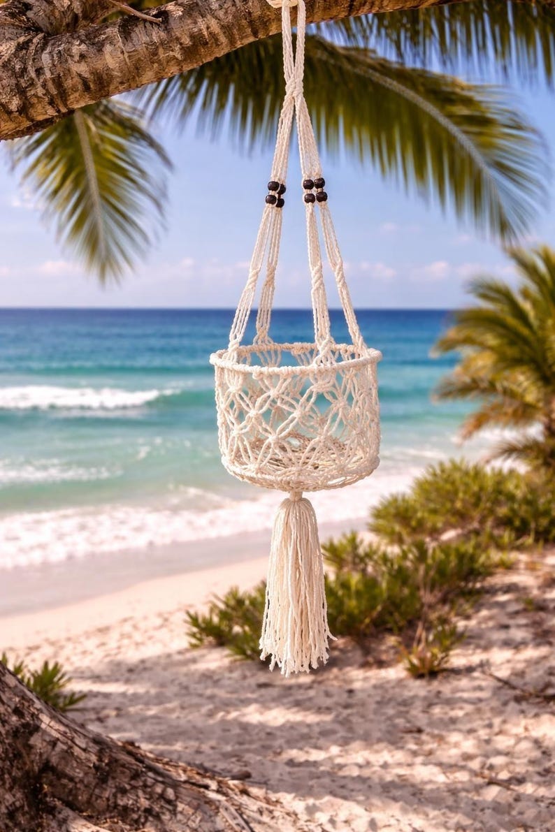 Pu&ograve; includere: Un portavaso in macram&egrave; color crema con un design a cestino intrecciato e una lunga nappa. Il portavaso &egrave; sospeso a un ramo d'albero sullo sfondo di una spiaggia con acqua blu e palme.