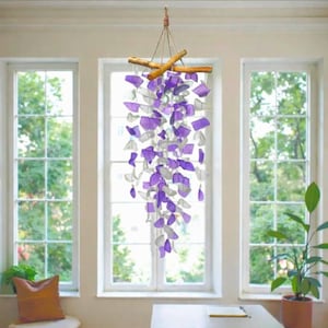 Puede incluir: Un móvil colgante con numerosas piezas de vidrio translúcido en tonos violeta y transparente, suspendidas de una rama de madera horizontal mediante hilos finos. El objeto decorativo muestra una disposición en cascada de formas geométricas.