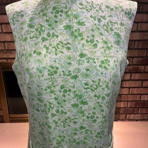 May include: Sleeveless dress with a white background and a green floral pattern. The pattern includes the words "Clove", "Chicory", "Mustard", and "Ginger". The dress has two pockets and a high neckline.
