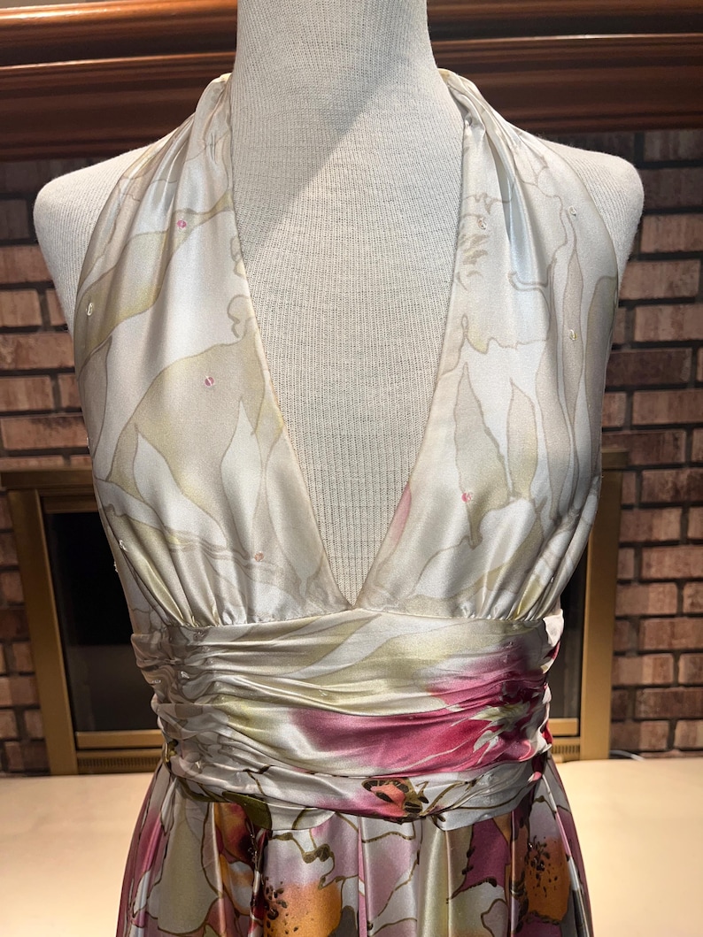 May include: A floral halter-neck dress. The dress has a V-neck and a gathered waist with a band of pink and red flowers. The fabric has a satin sheen. The dress is displayed on a mannequin, and the measurements are in centimetres.