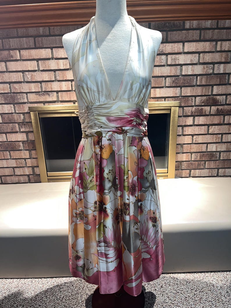 May include: A floral print halter dress on a mannequin. The dress has a V-neck, a gathered waist, and a knee-length skirt. The satin fabric displays a pattern of pink, orange, and white flowers. The dress is a vibrant piece.