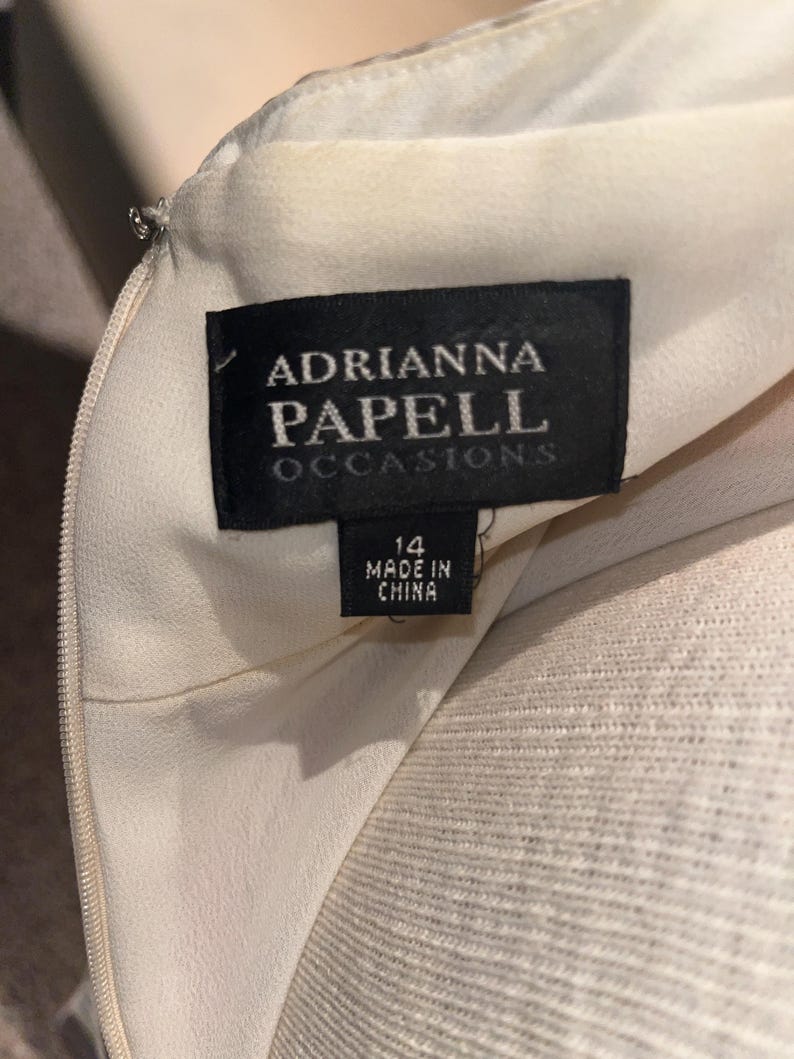 May include: Close-up of a cream dress with a black label reading "ADRIANNA PAPELL OCCASIONS." The label states the dress is size 14 and made in China. A silver zip is also visible.