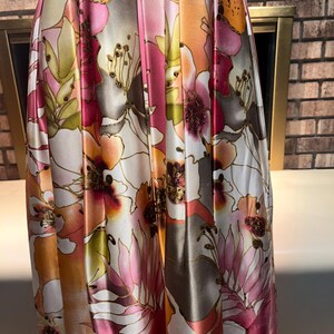 May include: A floor-length dress with a vibrant floral print in pink, orange, and white. The satin fabric catches the light, highlighting the large, stylized flowers with gold detailing. The dress has soft folds and drapes elegantly.