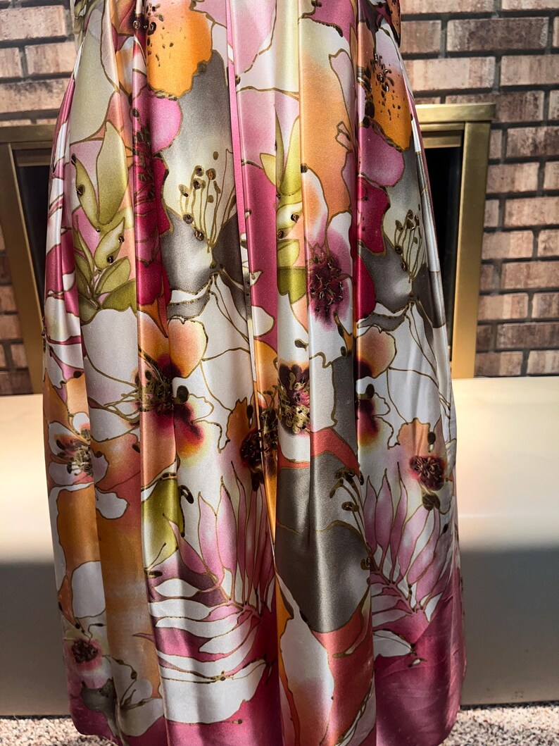 May include: A floor-length dress with a vibrant floral print in pink, orange, and white. The satin fabric catches the light, highlighting the large, stylized flowers with gold detailing. The dress has soft folds and drapes elegantly.