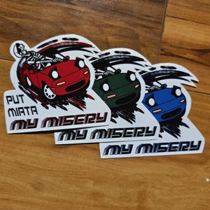 May include: A collection of die-cut stickers featuring stylized illustrations of a red, green, and blue convertible car. Each sticker has the text "PUT MIATA MY MISERY" in a bold font, with a skeleton driving the red car.
