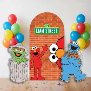 Peut inclure: Décoration de fête sur le thème de Sesame Street avec des découpes en carton d'Elmo, Cookie Monster et Oscar le Grognon. Une toile de fond en briques avec un panneau "Liam Street" est au centre, avec des ballons rouges, bleus, jaunes et verts.