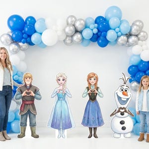 May include: A party scene featuring a balloon arch in shades of blue, silver, and white. Cardboard cutouts of characters from the movie Frozen are displayed in front of the arch, along with a young girl and a woman.
