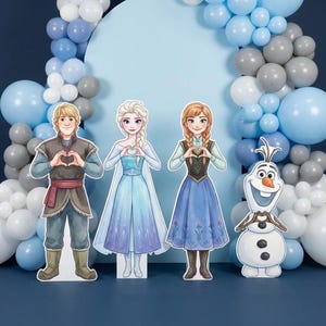 May include: Cardboard cutouts of characters from the animated film Frozen, including Kristoff, Elsa, Anna, and Olaf, are arranged in front of a blue backdrop with a balloon arch. The characters are holding their hands in a heart shape.