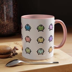 May include: White ceramic mug with a pink interior and handle. The mug features a repeating pattern of pixelated, colourful, cartoon-like characters. A spoon and a macaron are in the foreground, resting on a wooden surface.