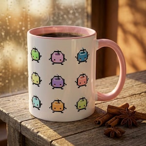 May include: White ceramic mug with a pink interior and handle, filled with coffee. The mug is decorated with pixelated, colorful fruit characters. Cinnamon sticks and star anise are arranged on a wooden surface next to the mug.