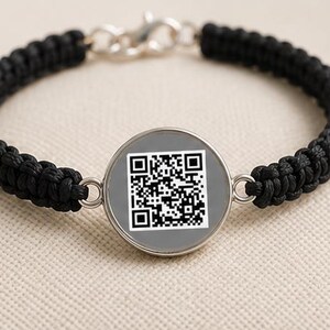 May include: A black woven cord bracelet featuring a central circular silver-toned charm. The charm displays a square black and white QR code printed on a gray background. The bracelet is secured with a silver-toned lobster claw clasp closure.