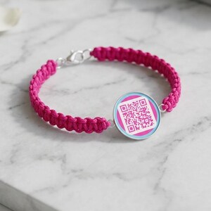 May include: A bright pink woven cord bracelet featuring a circular silver-toned charm. The charm displays a square QR code printed in pink against a white background, framed by a light blue border. The bracelet includes a silver-toned lobster clasp.