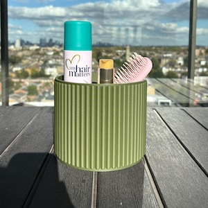 May include: A green, ribbed cylindrical container holds hair care products. A teal and white bottle with the text "my hair matters" is next to a tube of lip balm and a pink hairbrush. The container sits on a dark wooden surface.