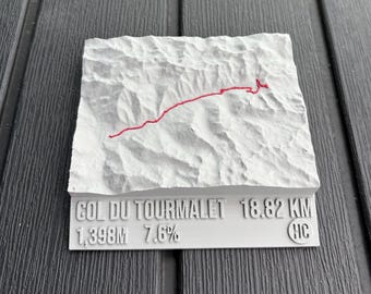 May include: A white, textured, rectangular object with a red line depicting a route. The text "COL DU TOURMALET 18.82 KM 1,398M 7.6%" is printed on the bottom. The object is on a dark grey surface.