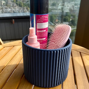 May include: A navy blue cylindrical container with vertical grooves holds hair care products. Items include a pink hairbrush, a pink spray bottle, and a black and blue hairspray can. The products are on a wooden table with a city view in the background.
