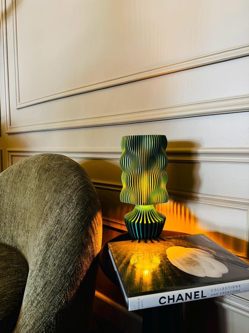 K&ouml;nnte beinhalten: Eine dekorative Lampe in Gr&uuml;n- und Gelbt&ouml;nen mit einem einzigartigen, geschichteten Design steht auf einem kleinen Tisch. Ein Buch mit dem Wort "CHANEL" ist zu sehen. Das Design der Lampe erzeugt ein welliges, strukturiertes Aussehen.