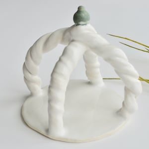 May include: White ceramic sculpture with a twisted rope design and a small green bird on top. The sculpture has a round base.
