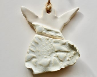 May include: A handmade ceramic angel ornament with outstretched wings. The angel is white with a textured, light blue-grey lower section. A natural twine loop is attached for hanging. Ideal for holiday decor or as a unique gift.
