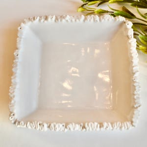 May include: A square, white ceramic dish with a ruffled edge. The dish has a slightly recessed center and a decorative, textured border. Greenery is visible in the upper right corner, providing a natural accent to the clean, white dish.