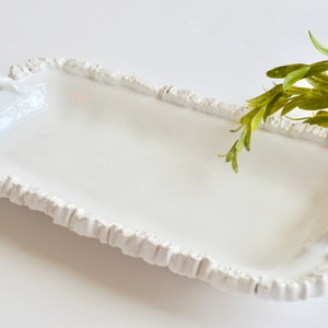 May include: White ceramic serving tray with rope-like handles on each side. The tray has a ruffled edge and is ideal for serving snacks or appetizers.
