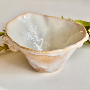 May include: A small, handmade ceramic bowl with a unique, organic shape. The interior is a glossy white, while the exterior features a blend of white and gold tones. The rim is accented with gold. Decorative greenery is in the background.