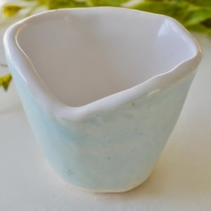 May include: A small, light blue ceramic bowl with a white interior. The bowl has an irregular, slightly rough texture.