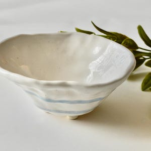 May include: A small white ceramic bowl with blue stripes around the rim. The bowl has an irregular, slightly wavy shape and sits on three small feet.