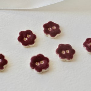 May include: Six flower-shaped buttons with a dark burgundy center and a white edge. Each button has two holes for sewing. The buttons are arranged on a white surface.