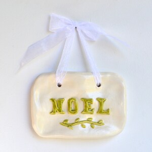 以下が含まれることがあります： 白いセラミック製のオーナメントで、緑色の「NOEL」の文字と緑色の葉の模様が描かれています。オーナメントは白いリボンで結ばれています。