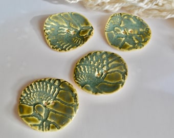 May include: Four handmade ceramic buttons. Each button is round, with a textured surface and a central hole for sewing. The buttons feature a blend of green and yellow-brown glazes, creating a natural, earthy aesthetic. Ideal for craft projects.
