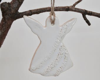 May include: A white ceramic angel ornament with outstretched wings, hanging from a natural twine string. The angel has textured details on the dress. The ornament is suspended from a brown tree branch against a white background. A decorative item.