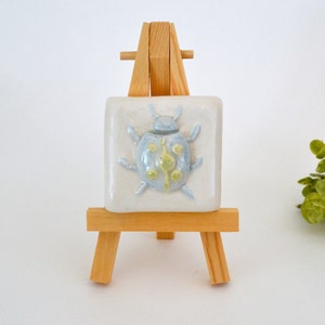 May include: A small ceramic tile with a blue and yellow ladybug design on a wooden easel.