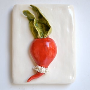 Puede incluir: Escultura de cerámica de un rábano rojo vibrante con hojas verdes sobre un fondo blanco texturizado. El rábano es el punto focal, mostrando un diseño detallado y artesanal. La obra de arte está montada sobre un fondo rectangular blanquecino.