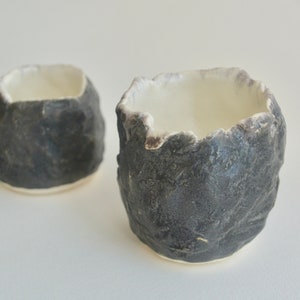 Puede incluir: Dos pequeñas tazas de cerámica negra con un exterior rugoso y texturizado y un interior esmaltado blanco. Las tazas son ligeramente diferentes en tamaño y forma.