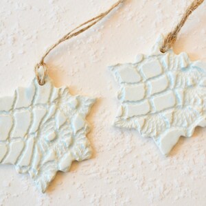 May include: Two white ceramic snowflake ornaments with a textured surface. Each ornament is attached to a piece of twine.