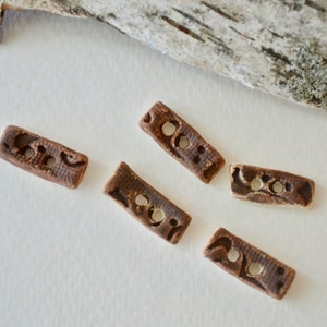 Puede incluir: Cinco botones de cerámica marrón con una superficie texturizada y un diseño decorativo. Los botones son de forma rectangular y tienen dos agujeros cada uno.