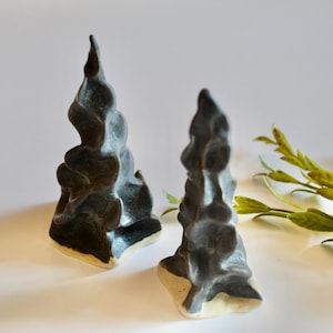 May include: Two black ceramic sculptures with a textured, swirling design. The bases are a light beige color. The sculptures are placed on a white surface with green leafy sprigs in the background. The sculptures have a unique, organic shape.