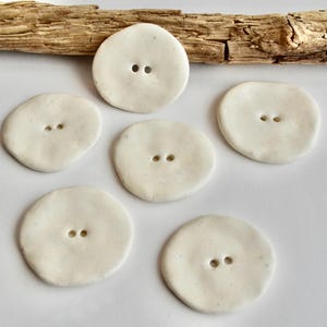 May include: A collection of six off-white ceramic buttons with two holes each. The buttons are round and oval-shaped, with a slightly textured surface. A piece of driftwood rests above the buttons on a white surface.