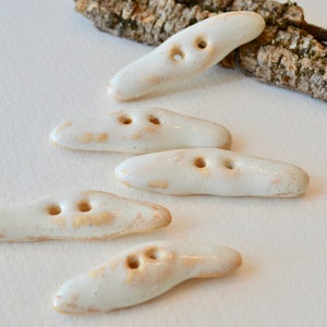 Pode incluir: Cinco botões de cerâmica branca com um esmalte marrom salpicado. Os botões têm a forma de ovais longos com dois furos cada.