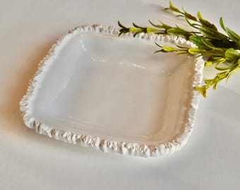 May include: A square, white ceramic dish with a textured, ruffled edge. The dish is empty and sits on a white surface. Green leafy sprigs are in the upper right corner.