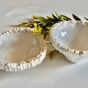 May include: Two white ceramic bowls with textured, ruffled edges. The bowls have a smooth interior and are placed on a white surface. Green and yellow sprigs of foliage are arranged around the bowls, adding a natural touch.