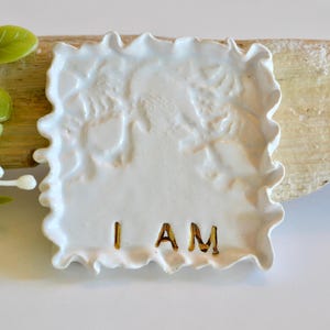 Puede incluir: Azulejo cuadrado de cerámica blanca con un diseño en relieve texturizado. El azulejo tiene un borde festoneado y las palabras "I AM" escritas en letras doradas.