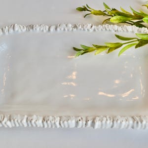 May include: A rectangular white ceramic serving tray with a ruffled edge. The tray is empty and has a glossy finish. Green leafy sprigs are placed on the top right corner of the tray. This tray is perfect for serving appetizers or desserts.